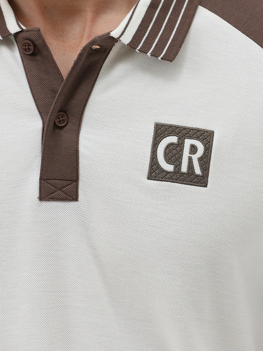 A man wearing a short-sleeve raglan polo shirt with a white body and brown sleeves, featuring a striped collar and a small 'CR' logo patch on the chest, paired with olive green pants against a light gray background.
