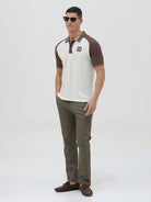 A man wearing a short-sleeve raglan polo shirt with a white body and brown sleeves, featuring a striped collar and a small 'CR' logo patch on the chest, paired with olive green pants against a light gray background.