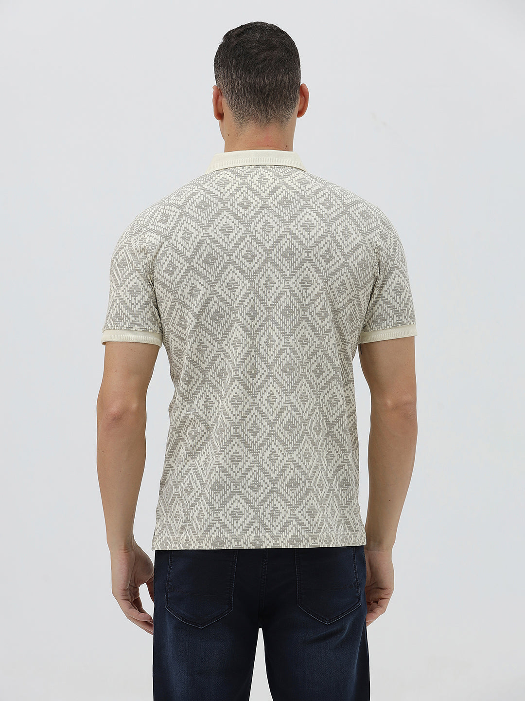 A man wearing a short-sleeve polo shirt with a beige and gray geometric pattern, paired with dark blue jeans, standing against a light gray background.