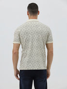 A man wearing a short-sleeve polo shirt with a beige and gray geometric pattern, paired with dark blue jeans, standing against a light gray background.