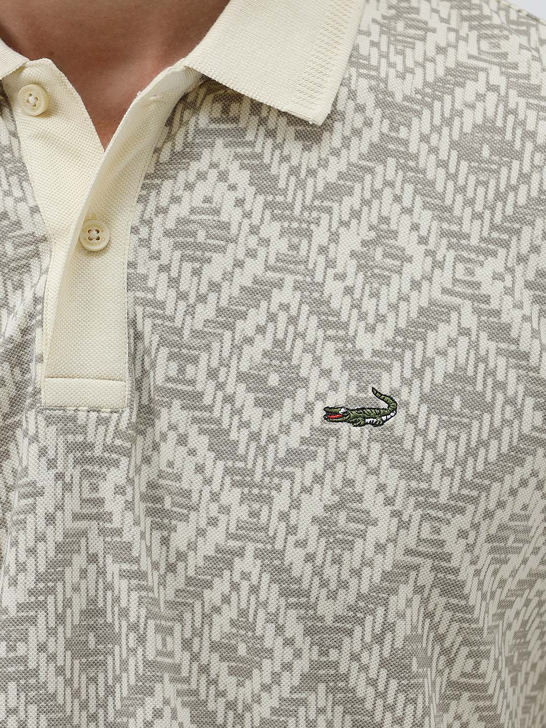 A man wearing a short-sleeve polo shirt with a beige and gray geometric pattern, paired with dark blue jeans, standing against a light gray background.