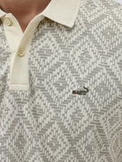 A man wearing a short-sleeve polo shirt with a beige and gray geometric pattern, paired with dark blue jeans, standing against a light gray background.