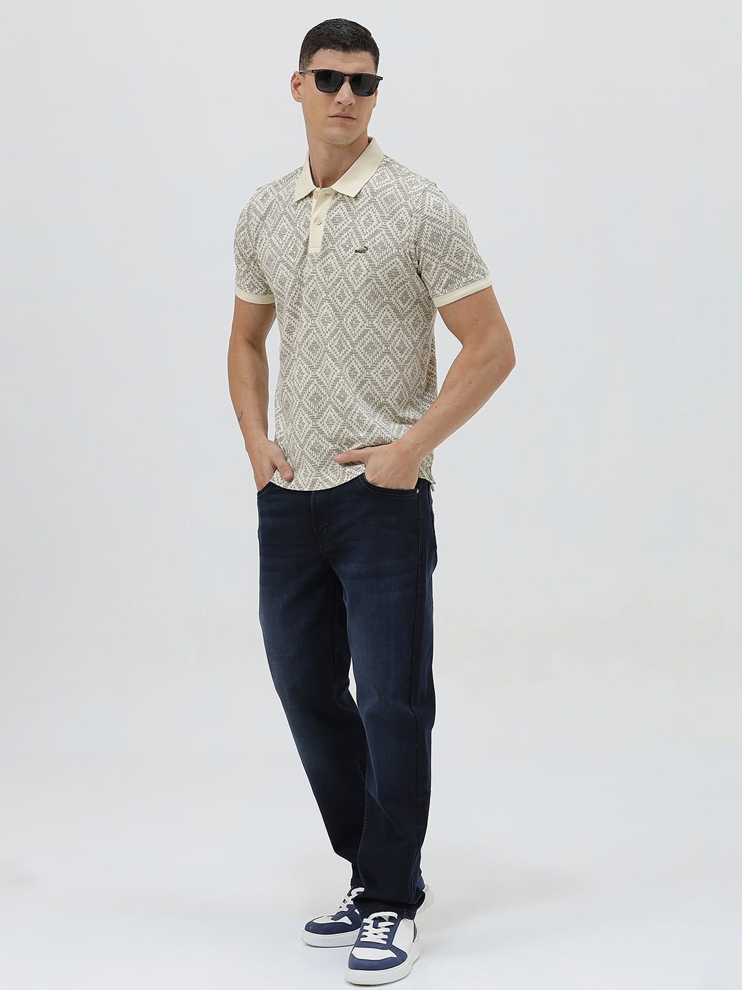 A man wearing a short-sleeve polo shirt with a beige and gray geometric pattern, paired with dark blue jeans, standing against a light gray background.
