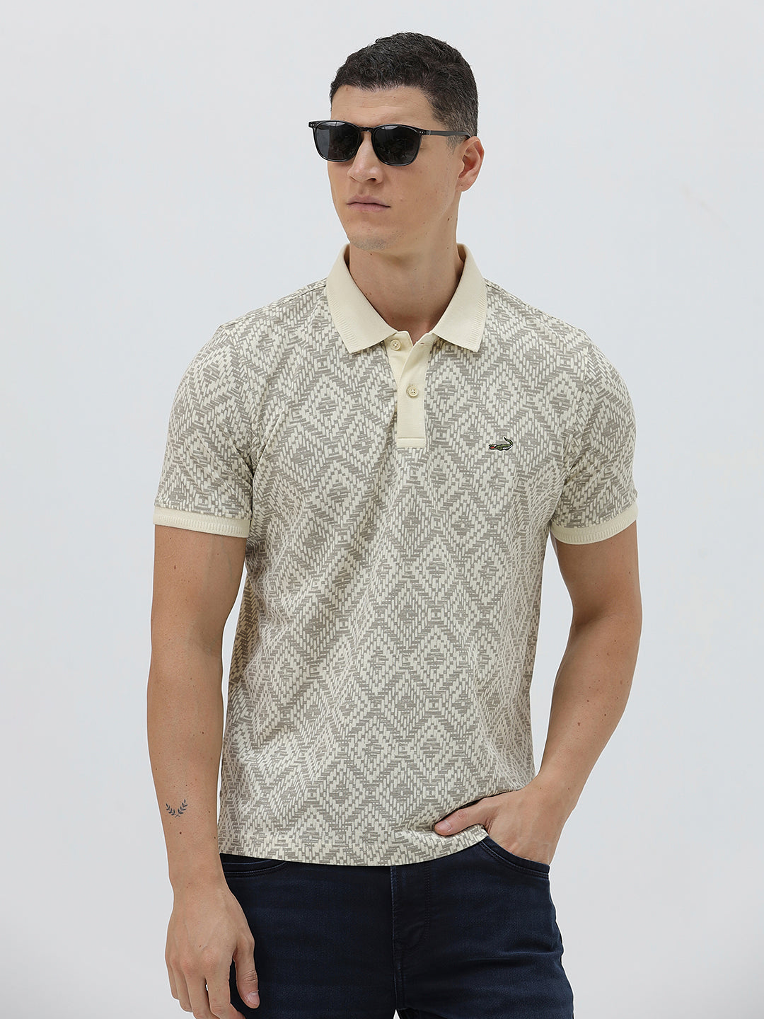 A man wearing a short-sleeve polo shirt with a beige and gray geometric pattern, paired with dark blue jeans, standing against a light gray background.