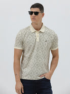 A man wearing a short-sleeve polo shirt with a beige and gray geometric pattern, paired with dark blue jeans, standing against a light gray background.