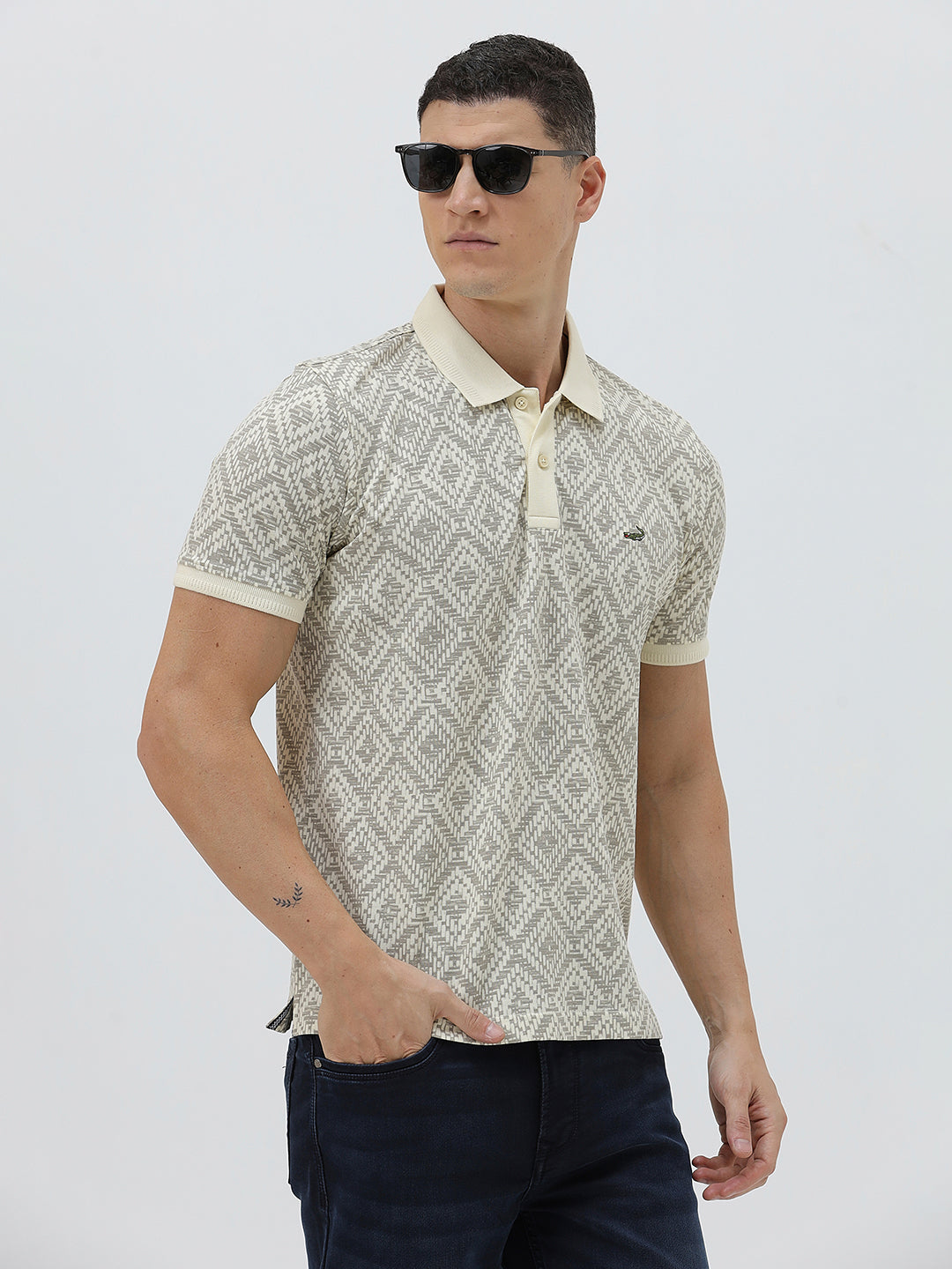 A man wearing a short-sleeve polo shirt with a beige and gray geometric pattern, paired with dark blue jeans, standing against a light gray background.