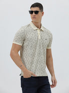 A man wearing a short-sleeve polo shirt with a beige and gray geometric pattern, paired with dark blue jeans, standing against a light gray background.