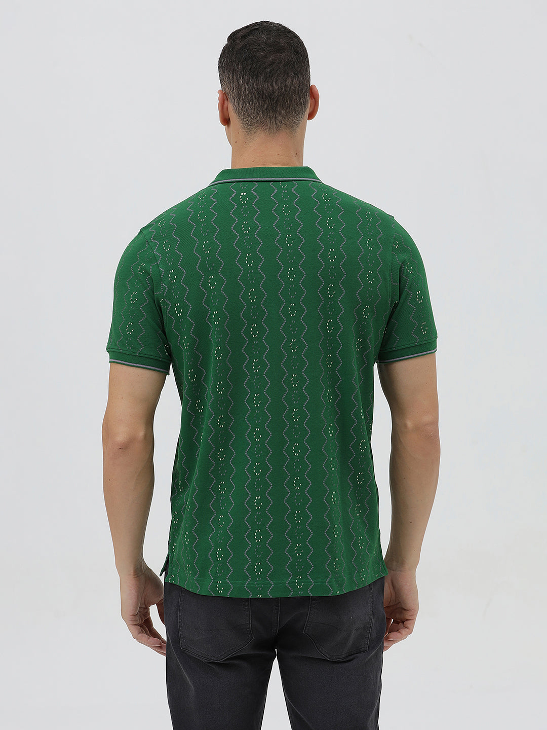 A man wearing a green short-sleeve polo shirt with a subtle geometric pattern, paired with dark pants and glasses, standing against a light gray background.