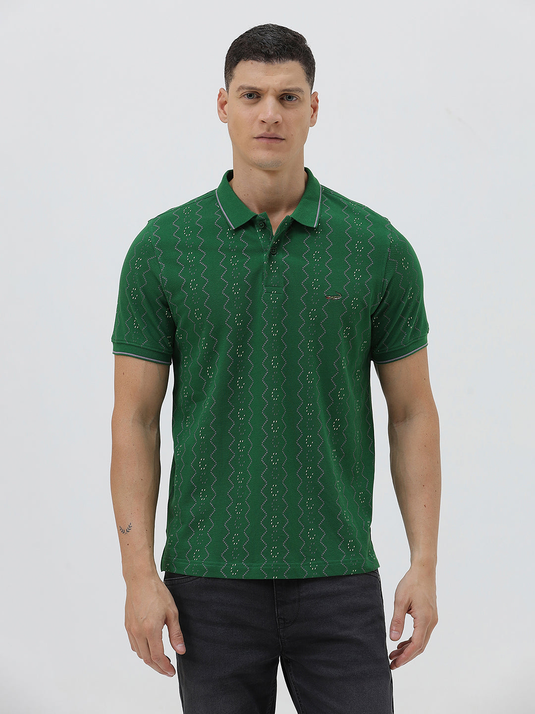A man wearing a green short-sleeve polo shirt with a subtle geometric pattern, paired with dark pants and glasses, standing against a light gray background.