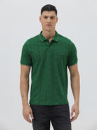 A man wearing a green short-sleeve polo shirt with a subtle geometric pattern, paired with dark pants and glasses, standing against a light gray background.