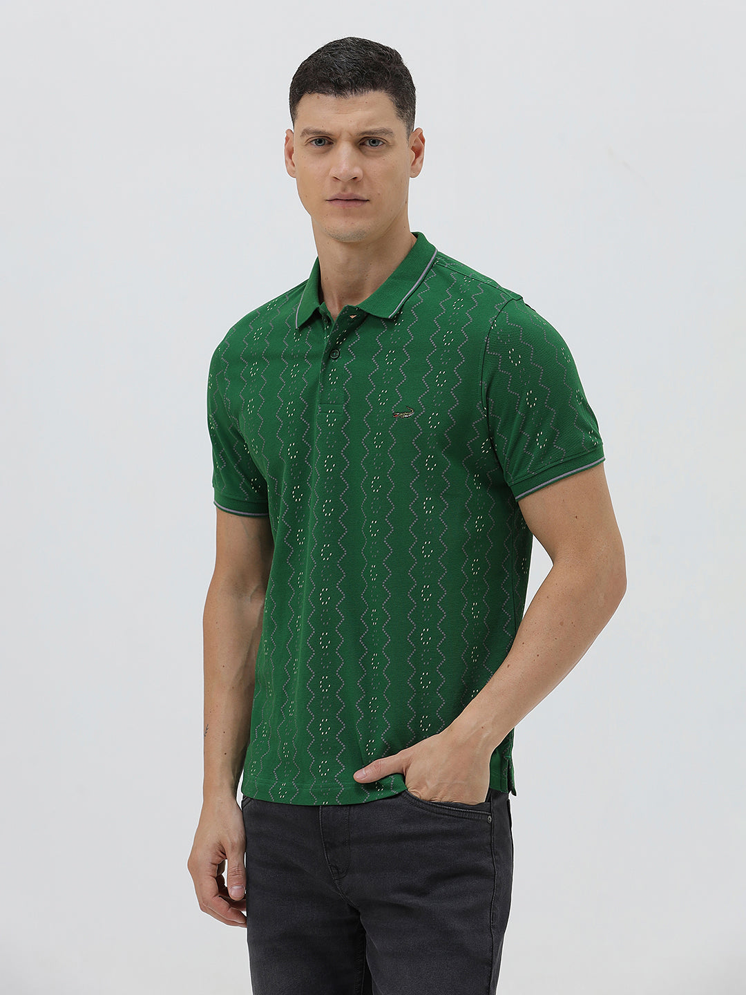 A man wearing a green short-sleeve polo shirt with a subtle geometric pattern, paired with dark pants and glasses, standing against a light gray background.