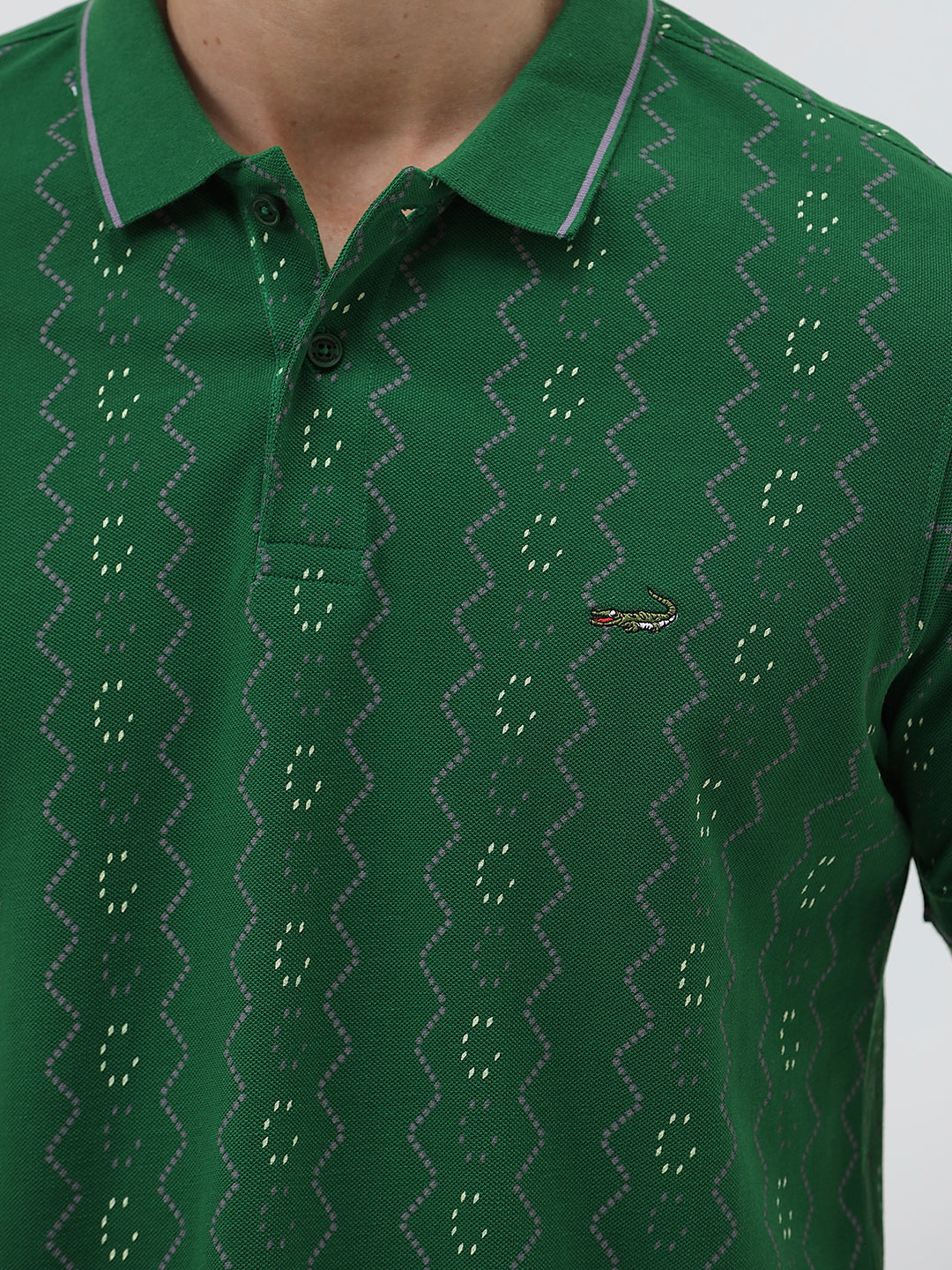 A man wearing a green short-sleeve polo shirt with a subtle geometric pattern, paired with dark pants and glasses, standing against a light gray background.