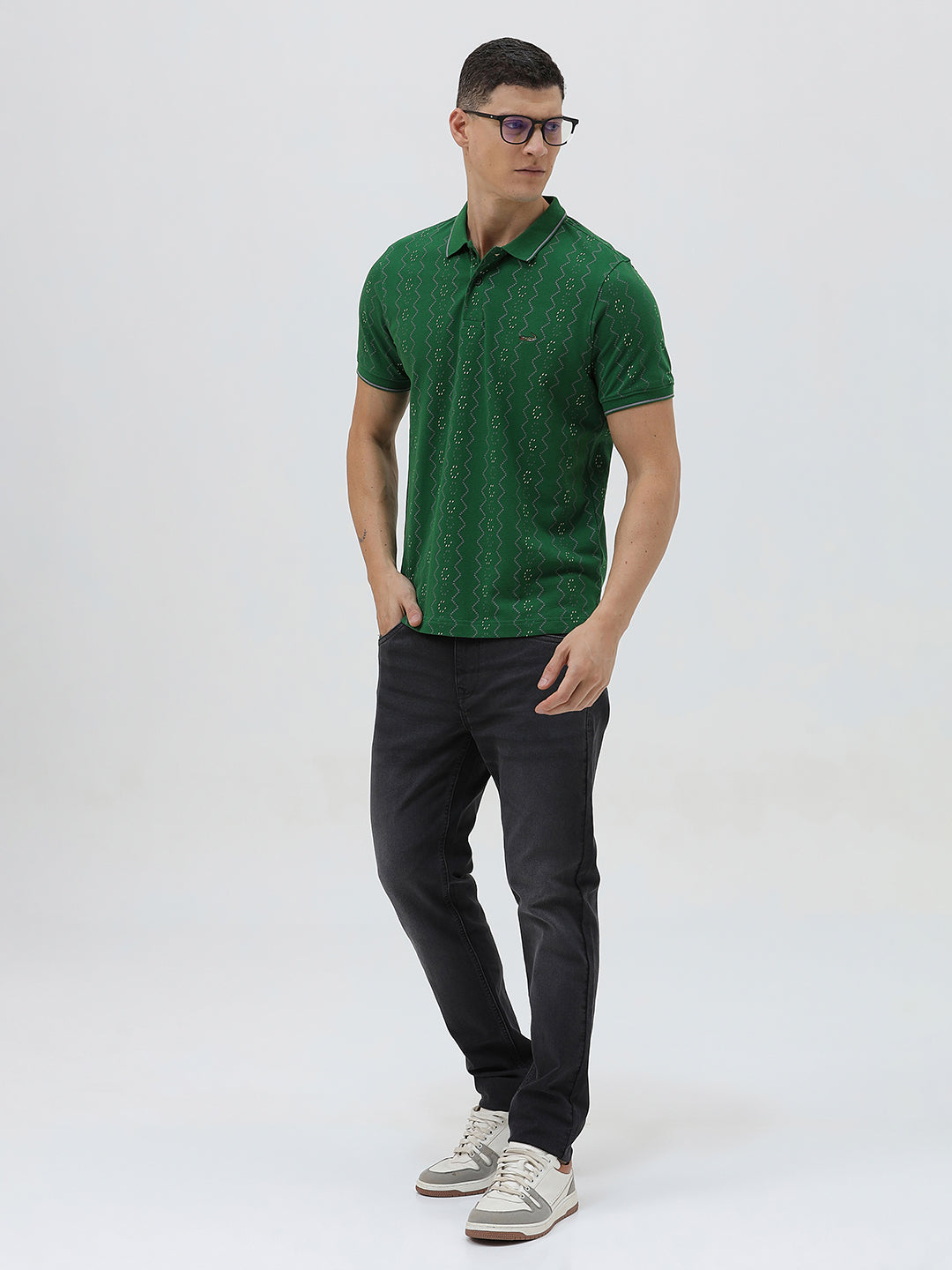 A man wearing a green short-sleeve polo shirt with a subtle geometric pattern, paired with dark pants and glasses, standing against a light gray background.