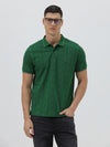 A man wearing a green short-sleeve polo shirt with a subtle geometric pattern, paired with dark pants and glasses, standing against a light gray background.