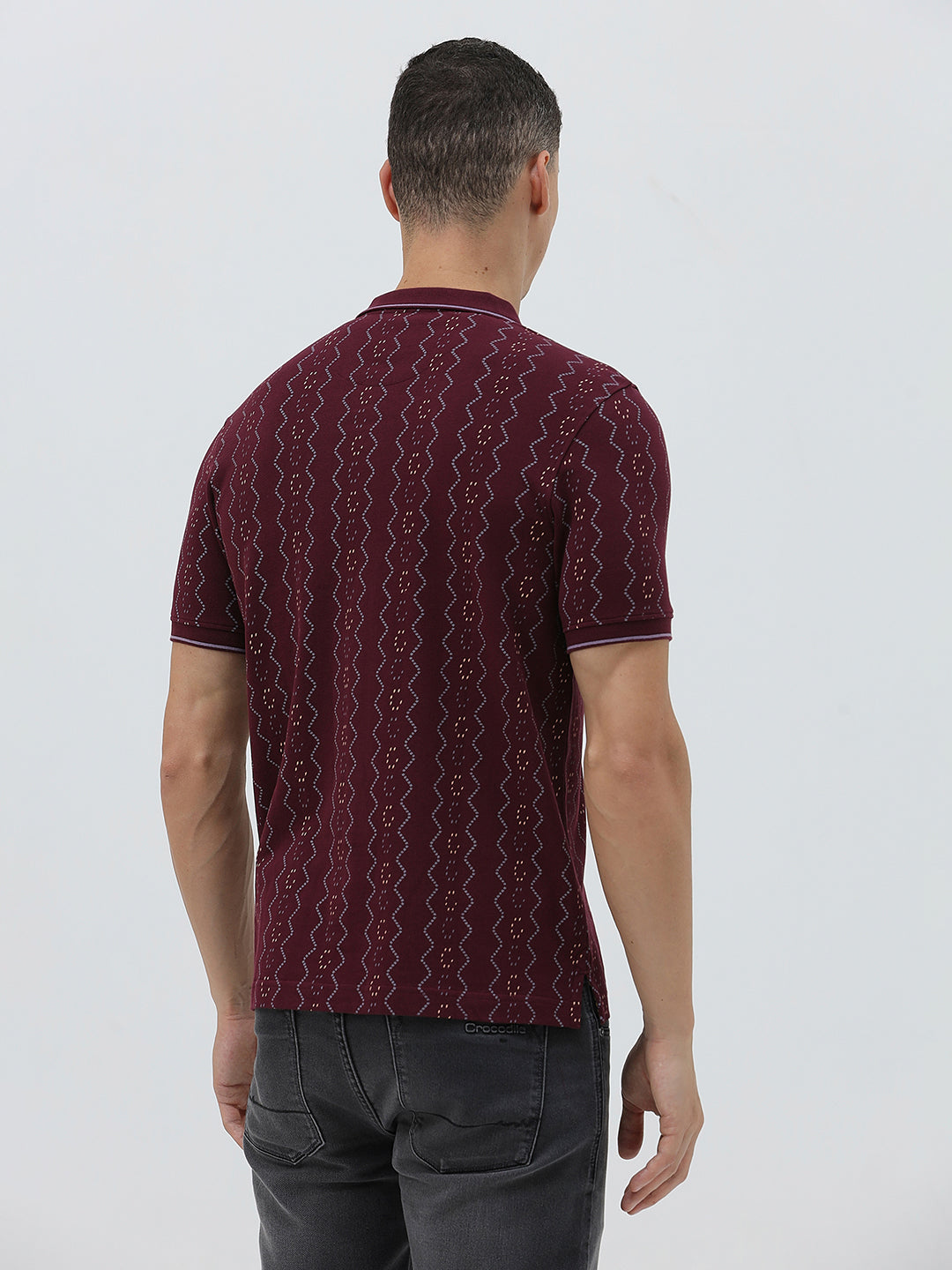 A man wearing a short-sleeve polo shirt in burgundy with a repeating vertical wavy pattern in dark and light red, paired with dark gray jeans, against a plain light gray background.