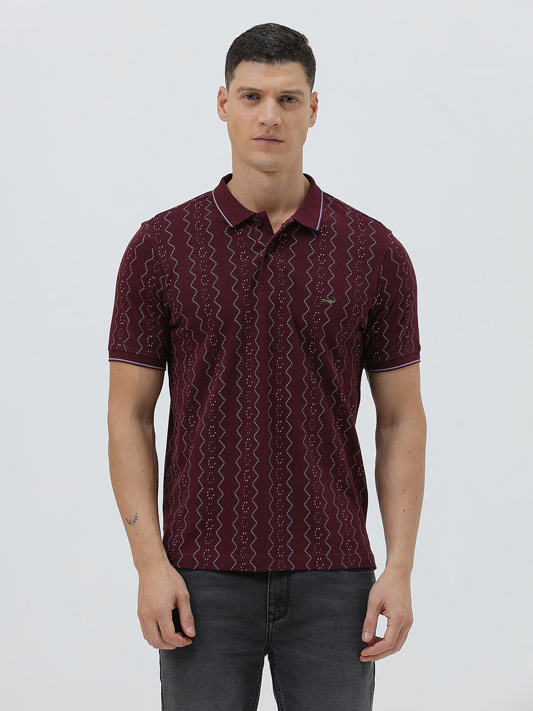 A man wearing a short-sleeve polo shirt in burgundy with a repeating vertical wavy pattern in dark and light red, paired with dark gray jeans, against a plain light gray background.