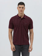 A man wearing a short-sleeve polo shirt in burgundy with a repeating vertical wavy pattern in dark and light red, paired with dark gray jeans, against a plain light gray background.