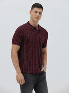 A man wearing a short-sleeve polo shirt in burgundy with a repeating vertical wavy pattern in dark and light red, paired with dark gray jeans, against a plain light gray background.