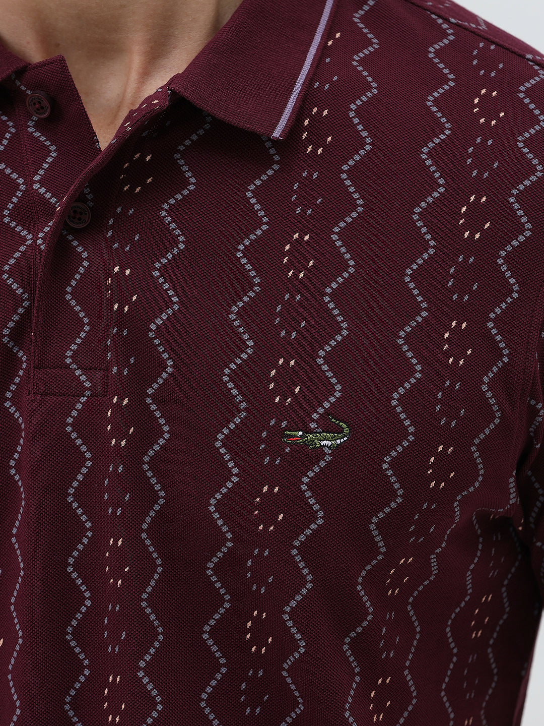 A man wearing a short-sleeve polo shirt in burgundy with a repeating vertical wavy pattern in dark and light red, paired with dark gray jeans, against a plain light gray background.