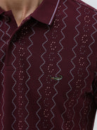 A man wearing a short-sleeve polo shirt in burgundy with a repeating vertical wavy pattern in dark and light red, paired with dark gray jeans, against a plain light gray background.