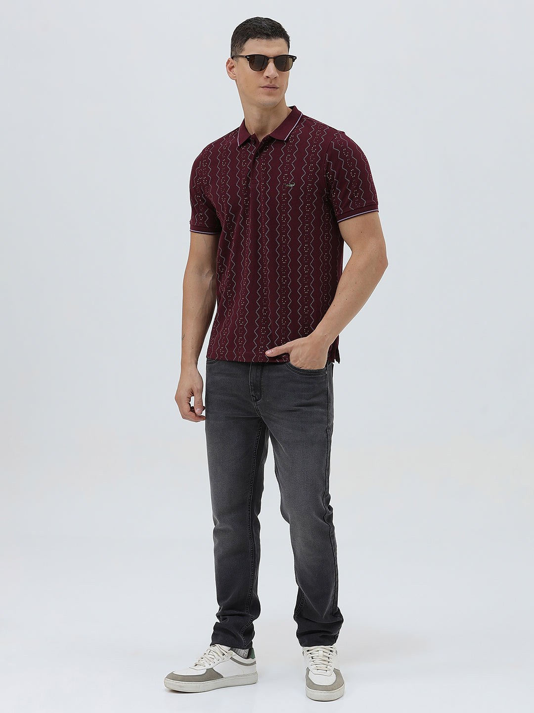 A man wearing a short-sleeve polo shirt in burgundy with a repeating vertical wavy pattern in dark and light red, paired with dark gray jeans, against a plain light gray background.