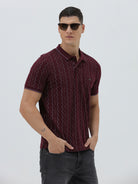 A man wearing a short-sleeve polo shirt in burgundy with a repeating vertical wavy pattern in dark and light red, paired with dark gray jeans, against a plain light gray background.
