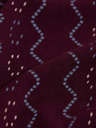 A man wearing a short-sleeve polo shirt in burgundy with a repeating vertical wavy pattern in dark and light red, paired with dark gray jeans, against a plain light gray background.