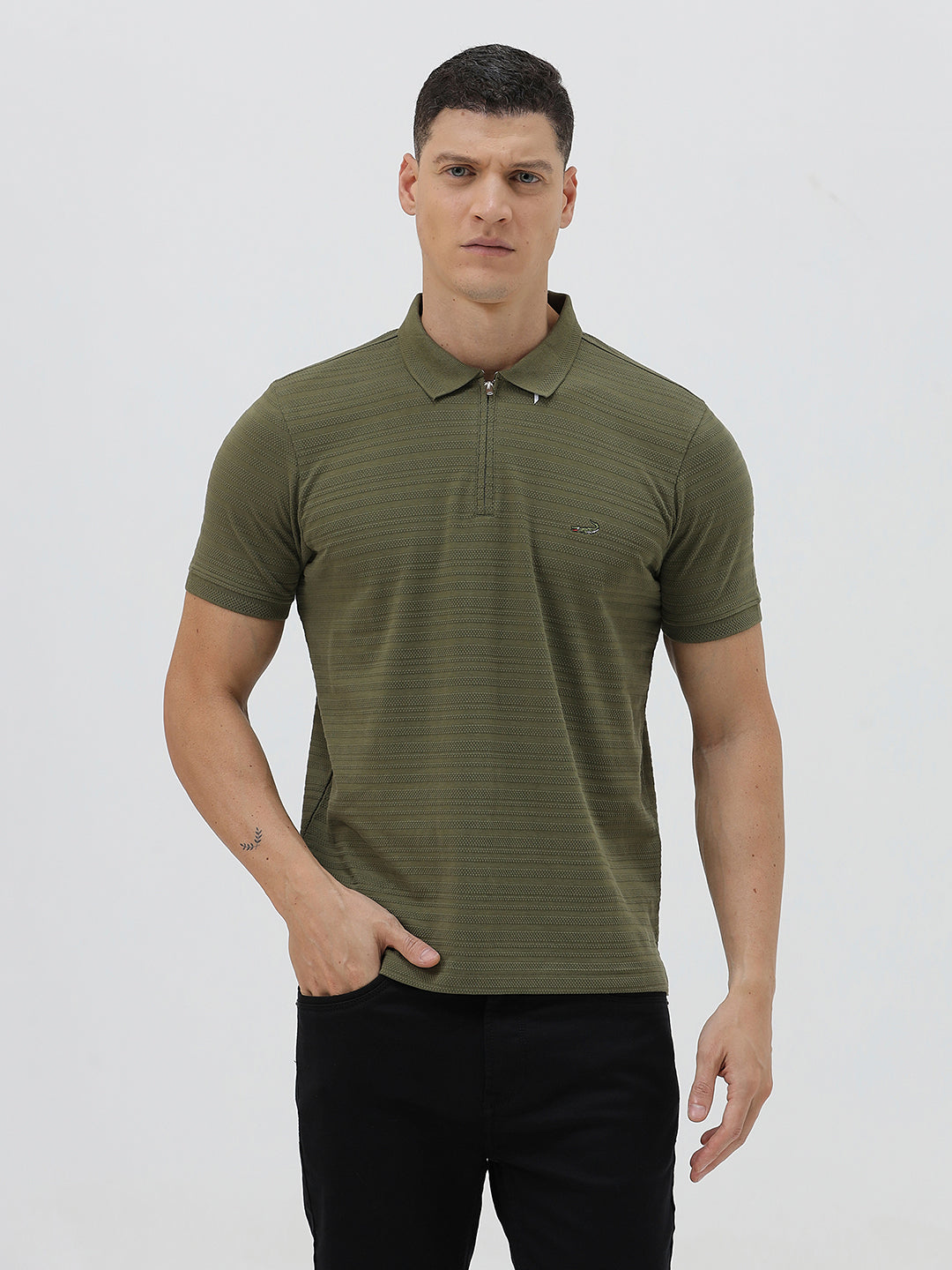 A man wearing an olive green short-sleeve polo shirt with a half-zip collar and black pants, standing against a light gray studio background.