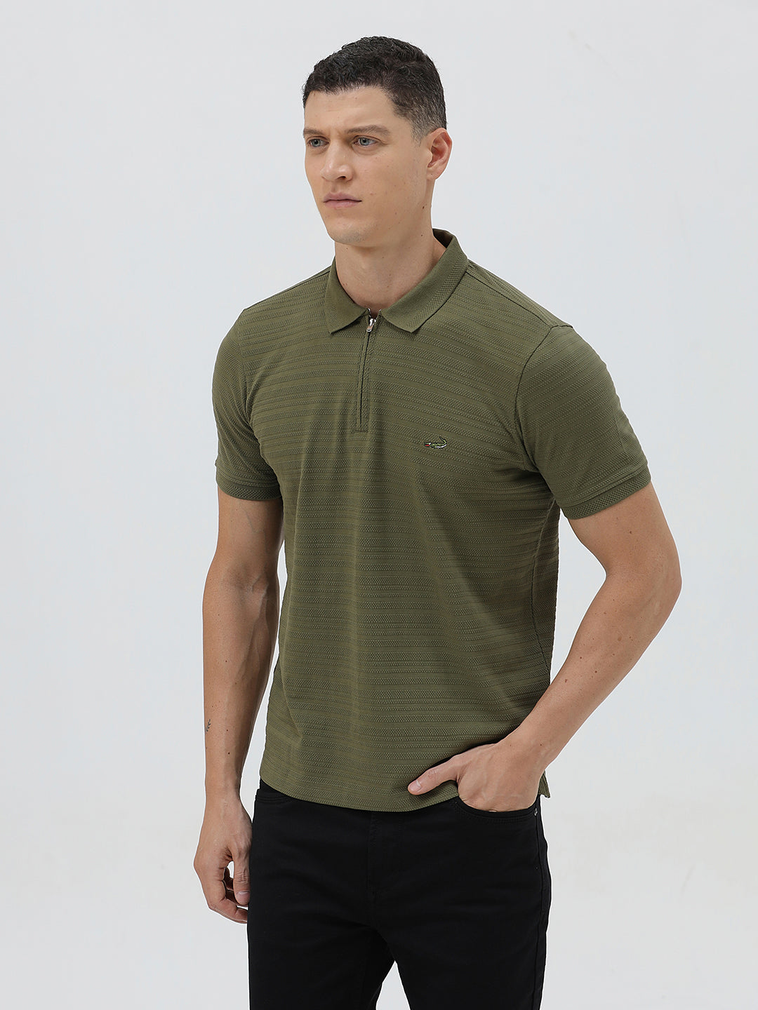 A man wearing an olive green short-sleeve polo shirt with a half-zip collar and black pants, standing against a light gray studio background.