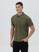 A man wearing an olive green short-sleeve polo shirt with a half-zip collar and black pants, standing against a light gray studio background.