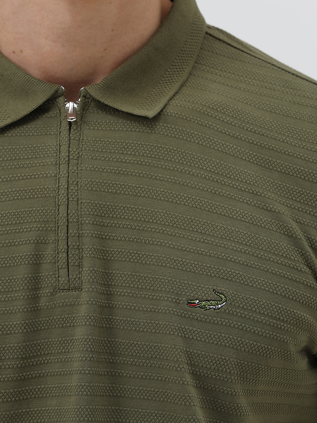 A man wearing an olive green short-sleeve polo shirt with a half-zip collar and black pants, standing against a light gray studio background.