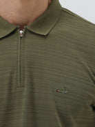 A man wearing an olive green short-sleeve polo shirt with a half-zip collar and black pants, standing against a light gray studio background.