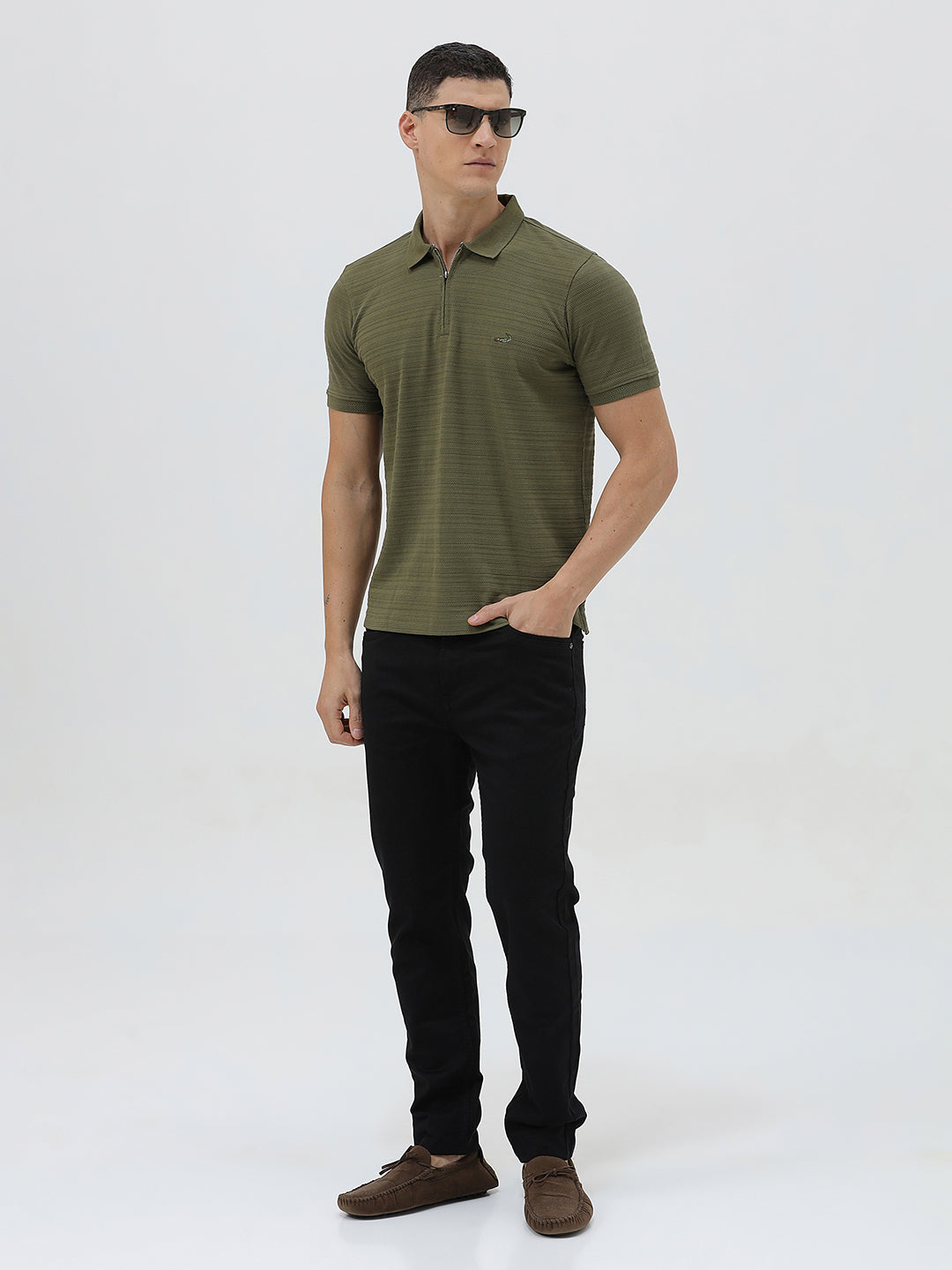 A man wearing an olive green short-sleeve polo shirt with a half-zip collar and black pants, standing against a light gray studio background.