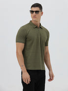 A man wearing an olive green short-sleeve polo shirt with a half-zip collar and black pants, standing against a light gray studio background.