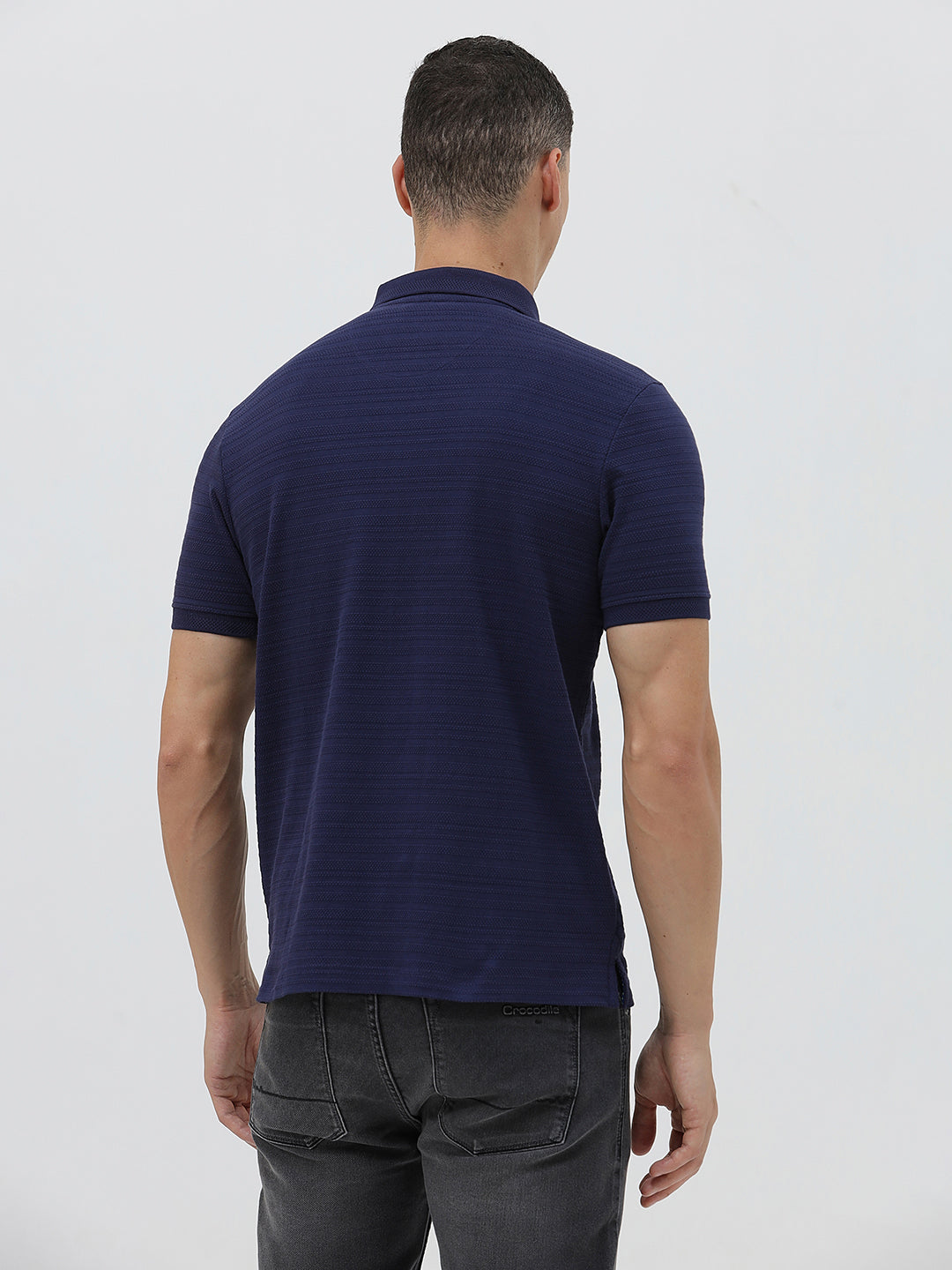 A man wearing a navy blue short-sleeve polo shirt with a small embroidered crocodile logo on the chest, paired with dark gray jeans, against a light gray studio background.