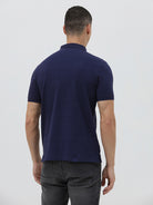 A man wearing a navy blue short-sleeve polo shirt with a small embroidered crocodile logo on the chest, paired with dark gray jeans, against a light gray studio background.