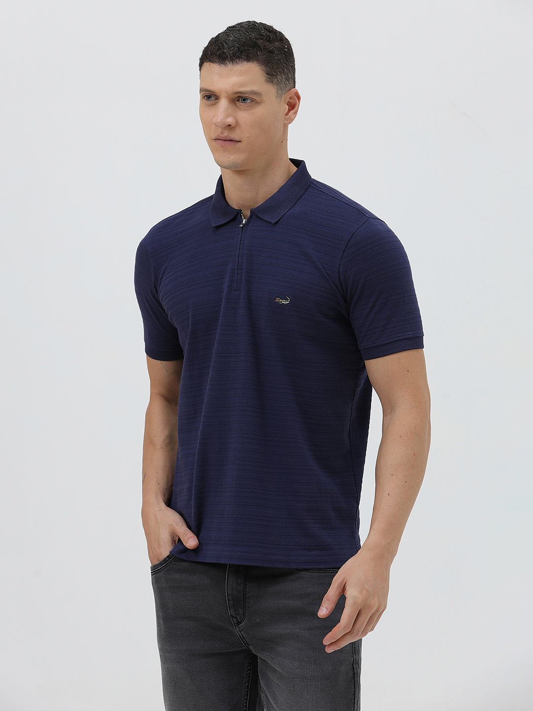 A man wearing a navy blue short-sleeve polo shirt with a small embroidered crocodile logo on the chest, paired with dark gray jeans, against a light gray studio background.