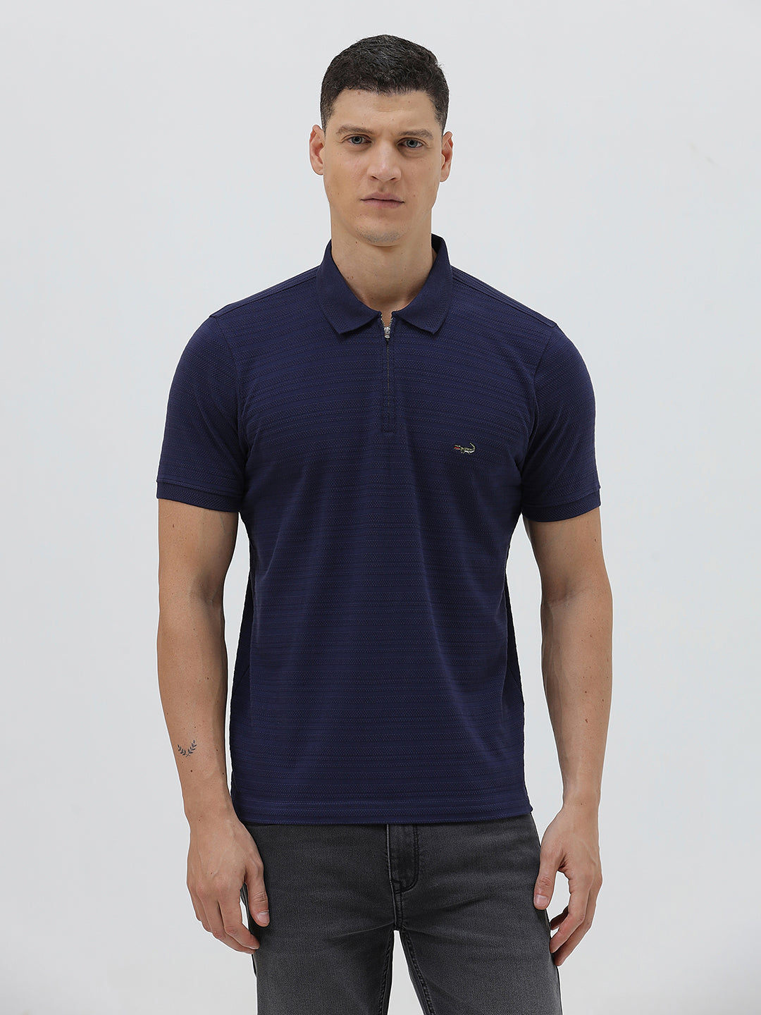A man wearing a navy blue short-sleeve polo shirt with a small embroidered crocodile logo on the chest, paired with dark gray jeans, against a light gray studio background.