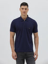 A man wearing a navy blue short-sleeve polo shirt with a small embroidered crocodile logo on the chest, paired with dark gray jeans, against a light gray studio background.