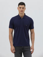 A man wearing a navy blue short-sleeve polo shirt with a small embroidered crocodile logo on the chest, paired with dark gray jeans, against a light gray studio background.