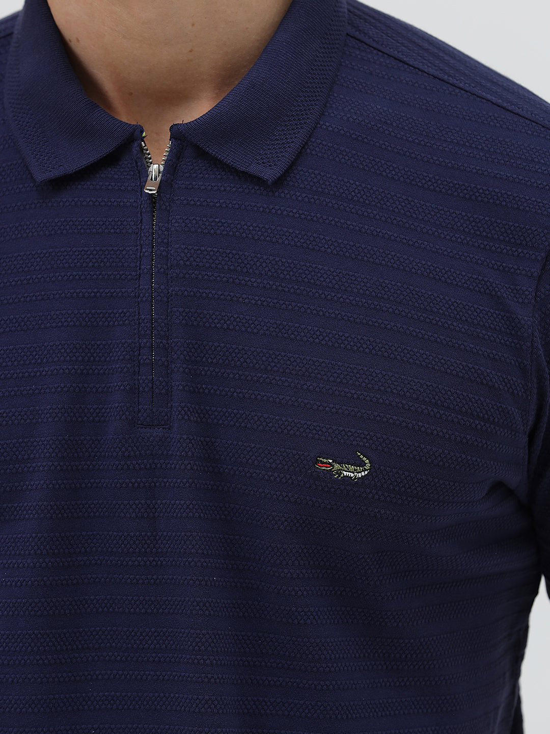 A man wearing a navy blue short-sleeve polo shirt with a small embroidered crocodile logo on the chest, paired with dark gray jeans, against a light gray studio background.