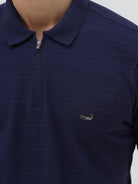 A man wearing a navy blue short-sleeve polo shirt with a small embroidered crocodile logo on the chest, paired with dark gray jeans, against a light gray studio background.