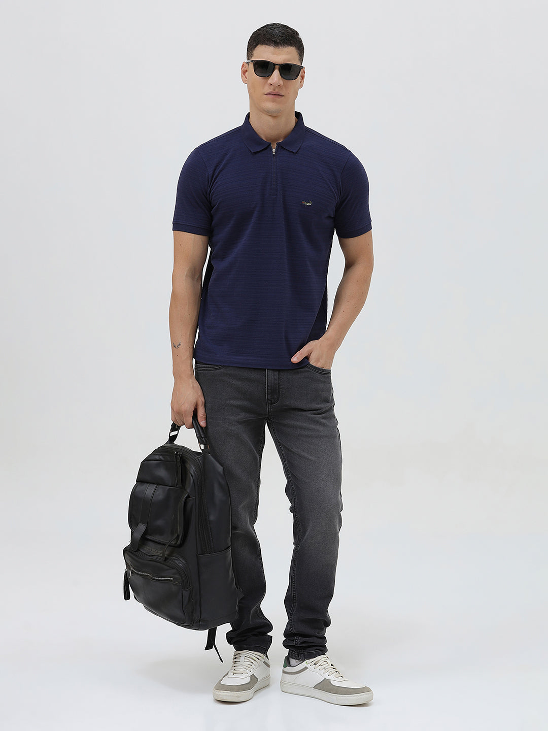A man wearing a navy blue short-sleeve polo shirt with a small embroidered crocodile logo on the chest, paired with dark gray jeans, against a light gray studio background.