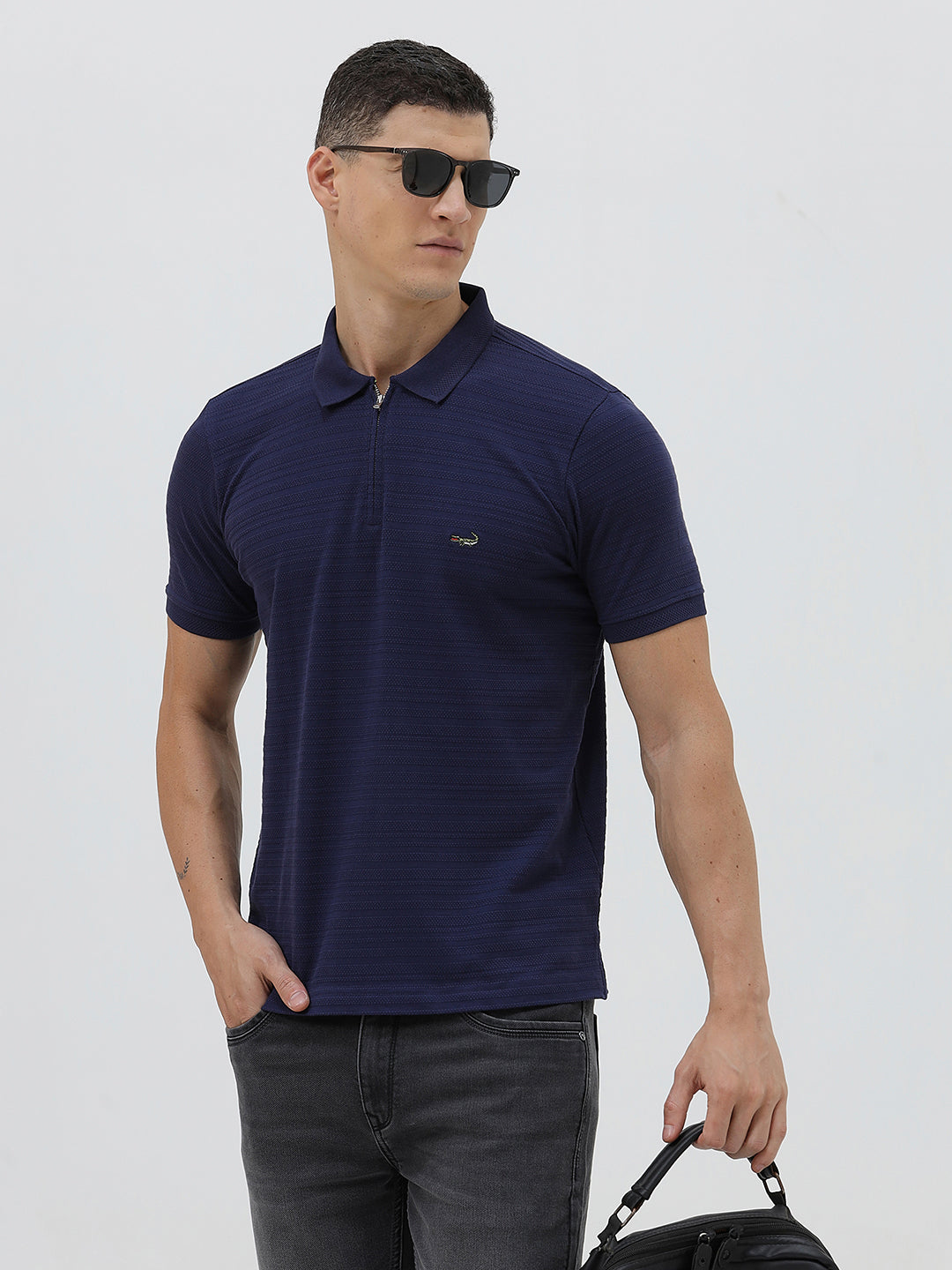 A man wearing a navy blue short-sleeve polo shirt with a small embroidered crocodile logo on the chest, paired with dark gray jeans, against a light gray studio background.