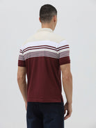 A man wearing a short-sleeve polo shirt with a colorblock design of cream, white, and burgundy, paired with dark blue jeans, against a plain light gray background.