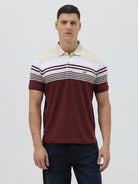 A man wearing a short-sleeve polo shirt with a colorblock design of cream, white, and burgundy, paired with dark blue jeans, against a plain light gray background.