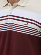 A man wearing a short-sleeve polo shirt with a colorblock design of cream, white, and burgundy, paired with dark blue jeans, against a plain light gray background.