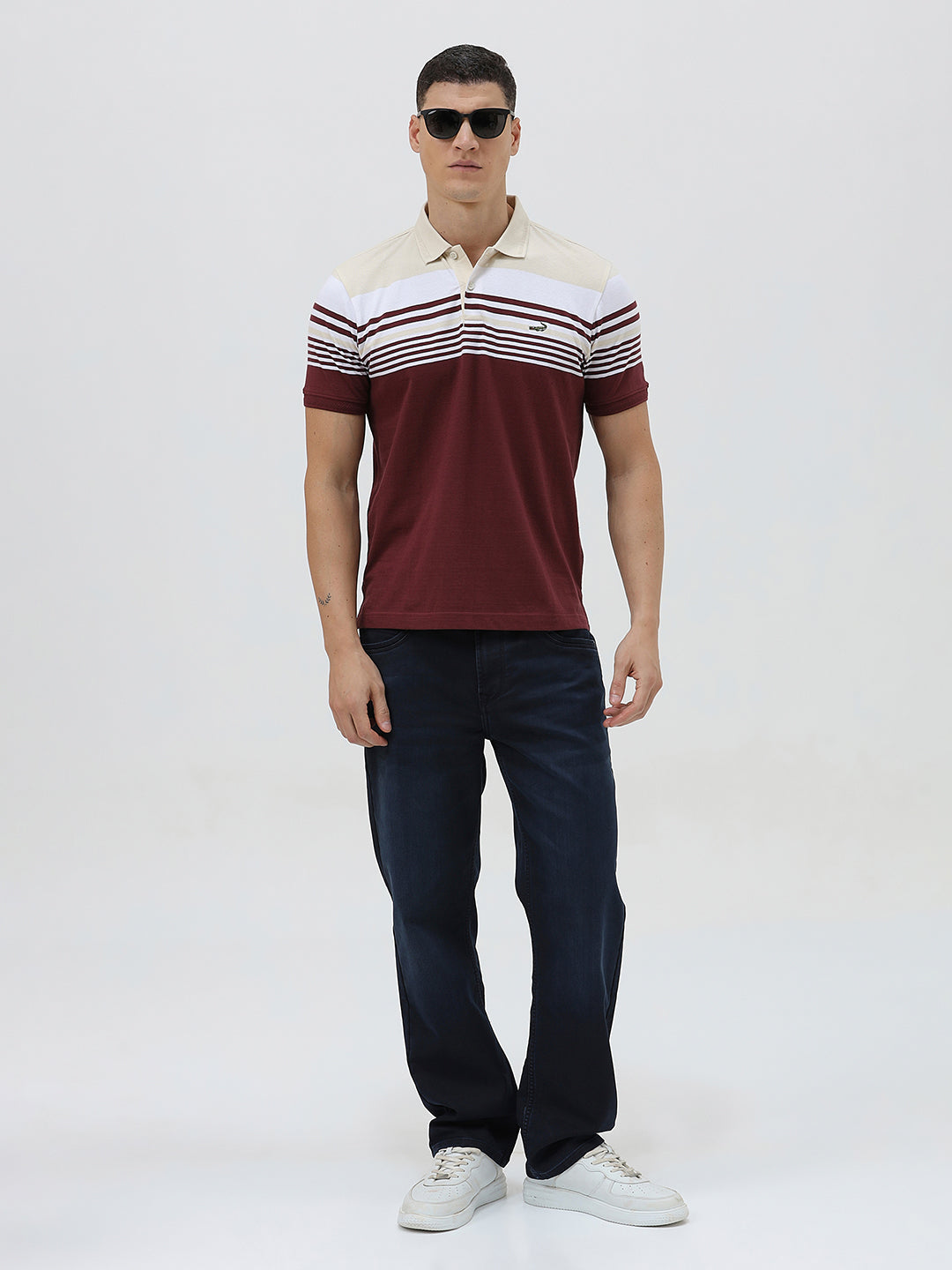A man wearing a short-sleeve polo shirt with a colorblock design of cream, white, and burgundy, paired with dark blue jeans, against a plain light gray background.