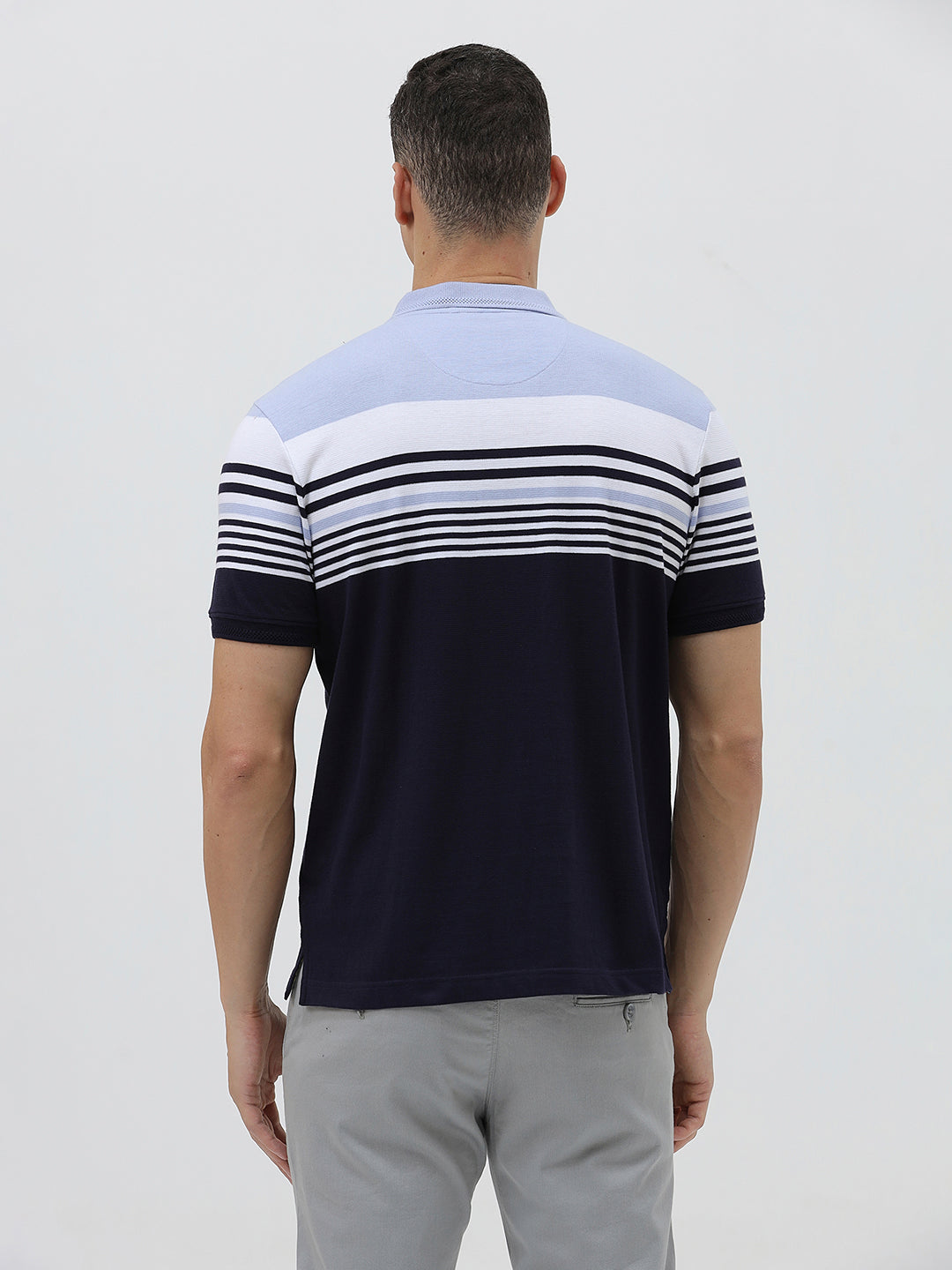 A man wearing a short-sleeve polo shirt with horizontal stripes in light blue, white, and navy, paired with light gray trousers, against a plain light gray background.