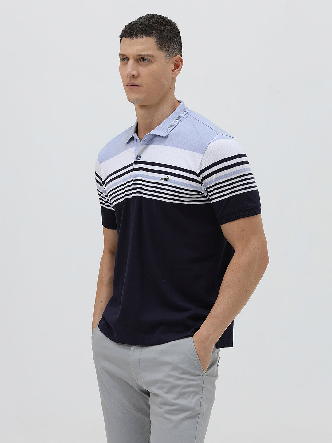 A man wearing a short-sleeve polo shirt with horizontal stripes in light blue, white, and navy, paired with light gray trousers, against a plain light gray background.
